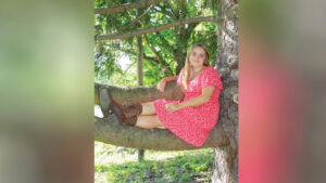 Nora Young, wearing a red dress with a small white repeating pattern and brown cowboy boots, sits on a large tree branch above the ground, resting one arm on another branch beside her. She looks at the camera with a soft smile. Trees with green leaves fill the background, with bright sunlight illuminating the ground beyond the shaded area where she sits.