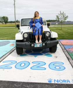 Nora Young sits on the hood of a white Jeep parked in a designated parking space painted with a blue and teal turtle and large blue “2026” lettering. The lower right corner features the handwritten name “NORA” with blue handprints on either side. Open fields, distant trees, houses and solar panels are visible in the background.