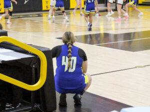 Nora Young squats at the side of a basketball court, watching the action during a game. She faces away from the camera and wears a blue and gold uniform over a black short-sleeve shirt, with a stylized “40” outlined in gold on the back of her jersey. Her long blonde hair is pulled into a bubble ponytail with multiple rounded sections.