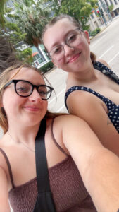 Two young women wearing sundresses and glasses pose for a selfie, each with a purse strap over her shoulder. Behind them are the glass windows and doors of a large building, palm trees, cars and a traffic light.