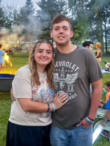 Nora Young poses with a young man, her arms around him and her hand resting on his stomach, as they both smile at the camera. Nora wears an American flag-themed shirt and matching nail art. Behind them, a person holds a sparkler while others gather at what appears to be a backyard Fourth of July celebration.