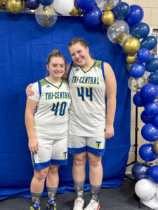 Nora Young poses with a friend and basketball teammate in front of a bright blue backdrop with a balloon arch in gold, clear and white. Both wear white high school basketball jerseys with blue lettering and yellow accents.