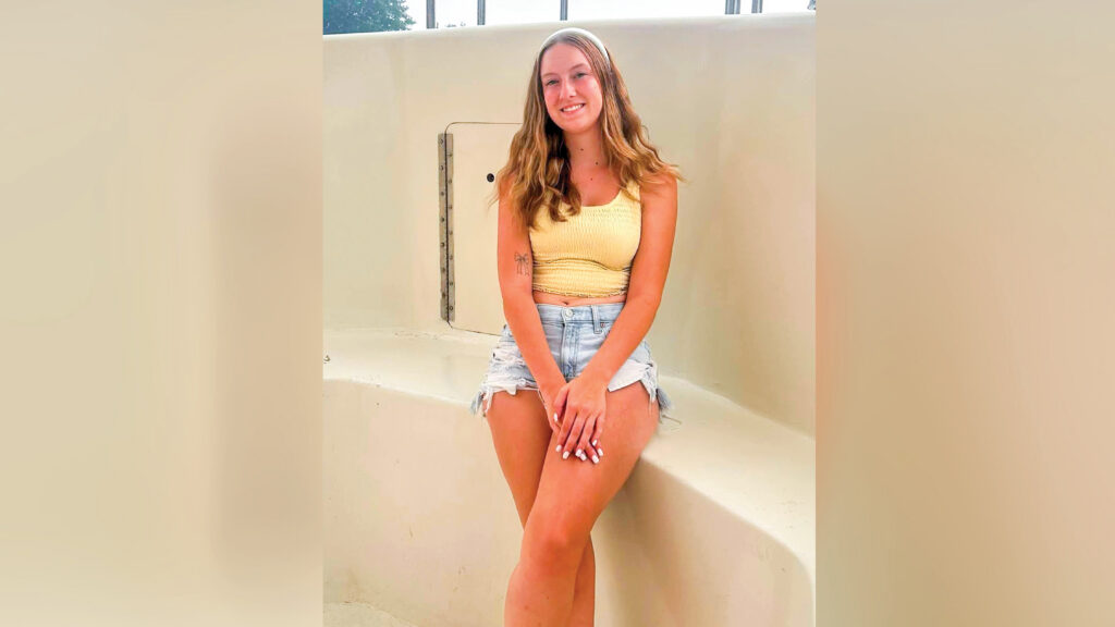 A smiling young woman with long, wavy hair and a white headband sits on the ledge of what appears to be an empty swimming pool. She wears a yellow smocked crop top and light blue denim shorts. Metal bars rise from the wall behind her, with a tree and overcast sky visible in the distance.