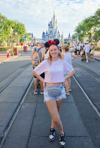 Kylee Bullock smiles with her hands on her hips and feet crossed while standing on a street at Disney World in Florida. She wears glittery Minnie Mouse ears with a shiny red bow, a pink Ball State crop top, light blue denim shorts, black-and-white sneakers, and a small gray waist bag. Other park visitors fill the street, with Cinderella Castle rising in the background.