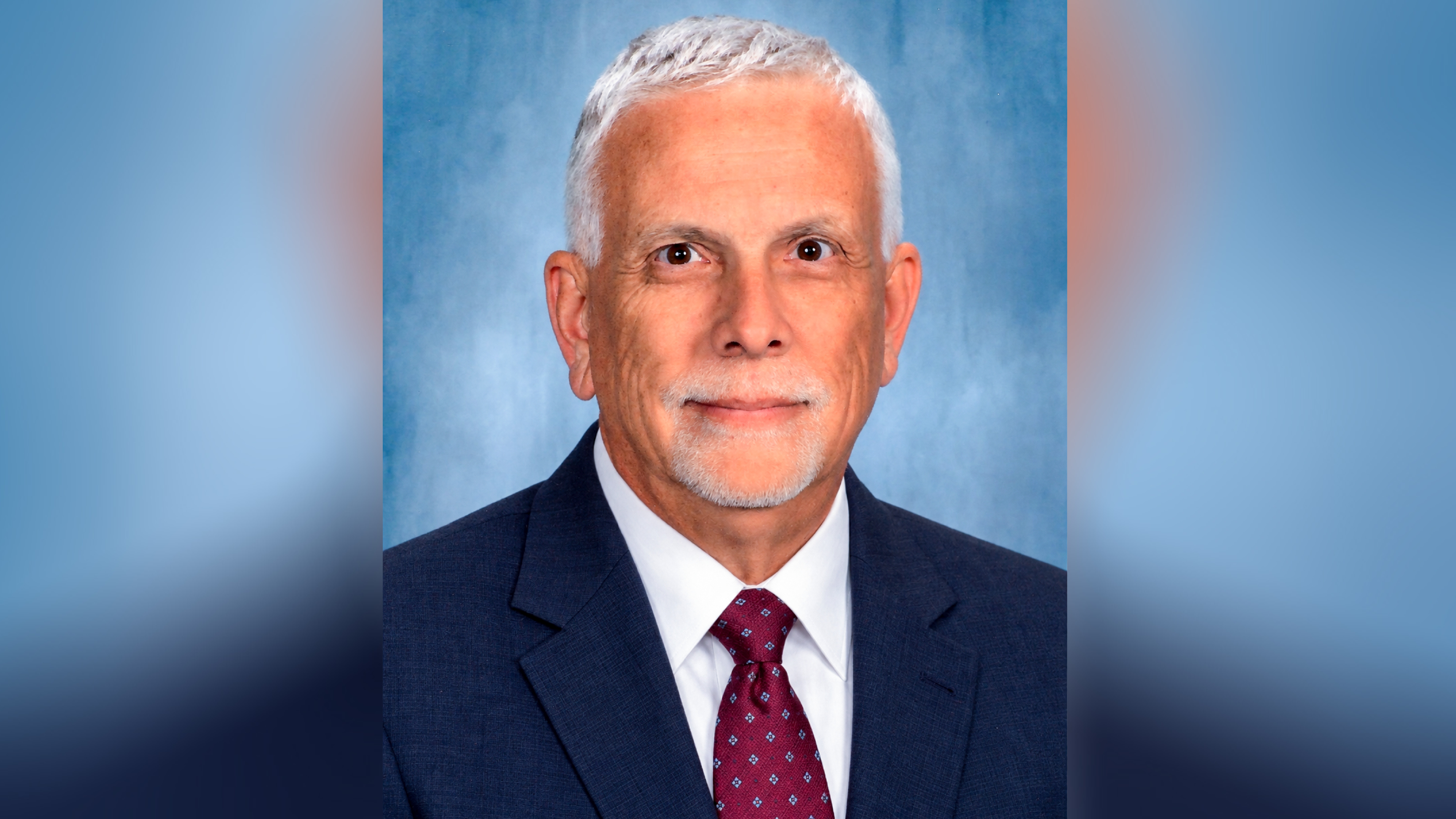 A professional headshot of an older man with short white hair, a goatee, and a mustache. He is wearing a navy-blue suit, white shirt, and patterned maroon necktie.