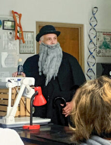 David Butler stands at the front of a science classroom dressed as Charles Darwin, wearing a black coat, bowler hat, and long gray beard and mustache. He holds an umbrella in one hand and a Charles Darwin bobblehead in the other. Science posters and a large model of a DNA strand are visible behind him.