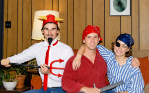 David Butler sits between two friends at a 1986 costume party. Butler and the young woman to his right are dressed as pirates, wearing bandanas and posing with a toy sword. The young man to his left is dressed as a devil with a horned hat, pointed goatee, red tie, and toy pitchfork. All three smile for the camera.