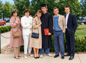David Butler poses outdoors with five people on his Ball State graduation day in 1989. He wears a black cap and gown and holds a red diploma cover with a gold Ball State insignia. Several people are bundled in coats, including a young woman standing close beside Butler.