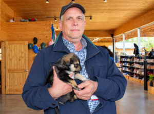 David Butler poses for a photo while holding a puppy inside a spacious, wood-paneled room at Happy Trails Kennels in Alaska. Shelves line one side of the room, with natural light coming through windows in the background.