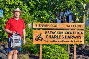 David Butler stands beside the welcome sign for the Charles Darwin Research Station on Santa Cruz Island in the Galápagos Islands. Wearing a red polo shirt, blue plaid shorts, and a tan hat, he holds a gray backpack and sunglasses. Lush greenery surrounds the sign.