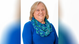 A headshot of a smiling woman with blonde hair, wearing a blue top and a patterned scarf in shades of blue, green, and white, against a white background.