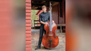 A young man in a black button-down shirt and black trousers smiles while standing on a stone patio and holding a large, polished wooden double bass. Behind him is a dark red, old-fashioned wooden house with a covered overhang. An opening in the building reveals a wooded area beyond.