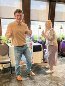 Marilynn Quick, wearing a silver top and lavender skirt, dances with a young man in a tan polo and jeans in a bright room decorated for a celebration.