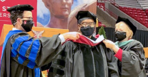 Marilynn Quick and another faculty member place a doctoral hood on a graduate during Ball State’s 2021 Commencement ceremony in Worthen Arena. All three wear doctoral regalia and face masks.