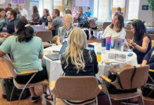 Marilynn Quick sits with five other women at a table as professionals use laptops and take notes during the 2022 Indiana School Counselor and Administrator Collaboration Institute at the Ball State Alumni Center.