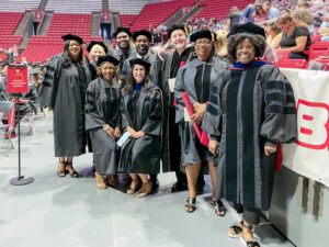 Educational Leadership faculty and graduating doctoral students pose together on the floor of Worthen Arena before Commencement. Wearing black doctoral gowns and tams, the group smiles for the camera, with Marilynn Quick in the back row, second from left. Other graduates and guests fill seats in the background.