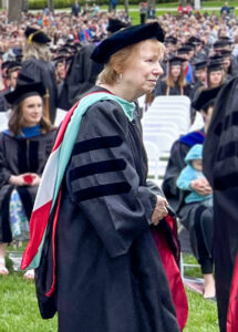 Marilynn Quick, wearing academic regalia and holding a doctoral hood, waits to participate in a hooding ceremony during Spring Commencement on the Ball State Quad. Graduates and guests are seated in rows behind her.