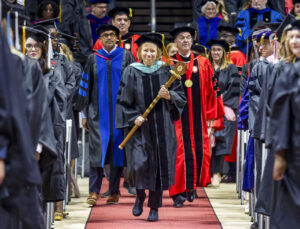 Marilynn Quick, wearing academic regalia, carries Ball State’s ceremonial mace down the center aisle of Worthen Arena during Commencement as graduates stand on either side.