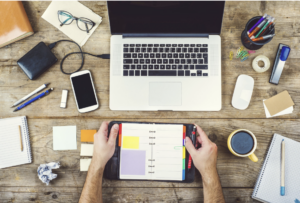 Overhead picture of a desktop with schedule, laptop, coffee, and other writing supplies.