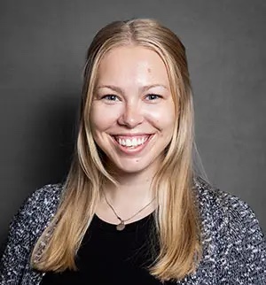A head-and-shoulders portrait of Mary Lowry, a woman with long blonde hair and a friendly smile. She is wearing a black top under a grey and white patterned cardigan and a simple pendant necklace. She is posed against a neutral, dark grey background.