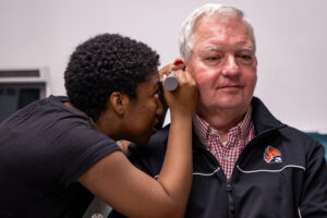 Adult Male with white hair, is having an exam from a student audiologist, with an Otoscope.
