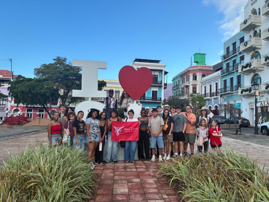 A group of Ball State Students stands in front of an I Heart San Juan statue in the heart of the city. The students in the middle hold a red flag with a white Ball State University Logo. You can see colorfully painted buildings on the left and right side of the photo, along with a bright blue sky. The students are smiling for the photo. Photo provided by Dr. Jean Marie Place.