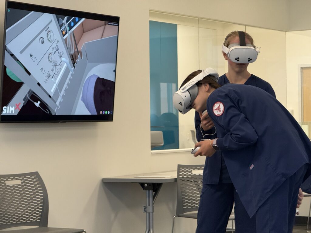The Scene is a School of Nursing Classroom, on the upper left is a televison screen depicting a 3-D model of a patient vitals screen readout. On the right hand side are two nursing students wearing virtual reality goggles covering their eyes, and they are holding controllers in their hands. They are interacting with the vitals screen pictured on the TV. 