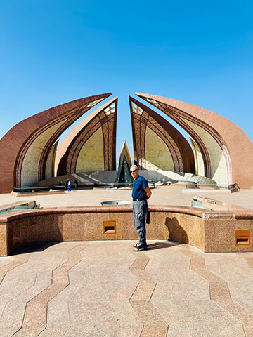 Dr. Larry Gerstein stands in front of the Pakistan Monument in Islamabad. It is a national landmark representing the country's unity, cultural diversity, and history. Completed in 2007 on Shakarparian Hill, the flower-shaped structure features four large petals (Punjab, Sindh, Balochistan, Pashtun) and three smaller ones (territories), surrounding a central star.