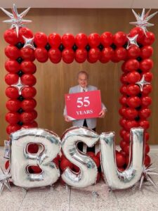 Dr. Ron Dolon stands under a red rectangular balloon arch, behind silver baloon letters spelling out BSU. He is holding a red sign that says 55 years, to celebrate his 55 years of service.