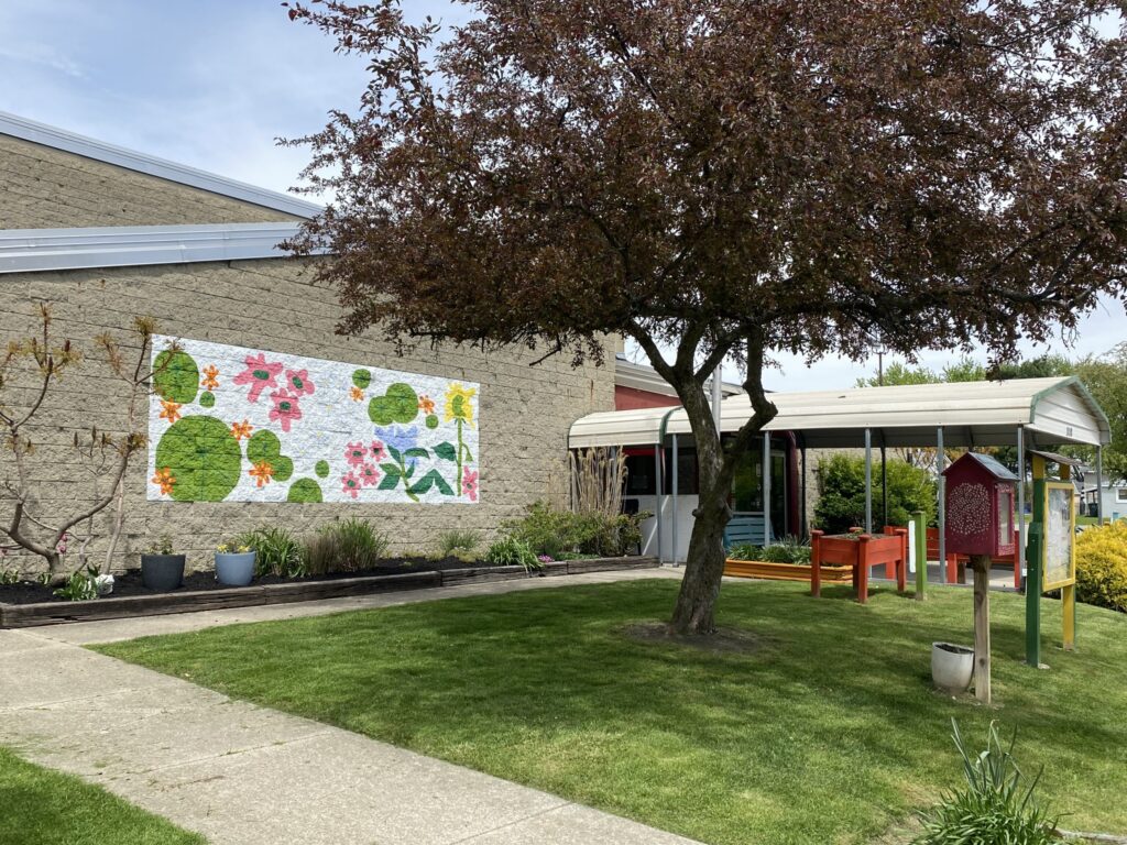 The exterior of the Ross Community Center Building. It is gray brick, with a colorful mural of flowers. Also seen in the shot, is a covered walkway leading to the entrance, and green landscaping including a tree, green grass, and several flower boxes. 