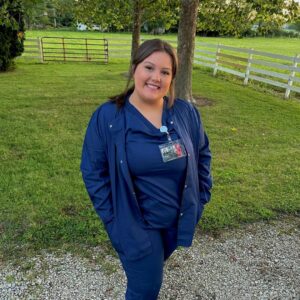 Nursing Student Aleyah, wearing blue scrubs, and standing with her back to a field of green grass, with a fence and trees in the background.