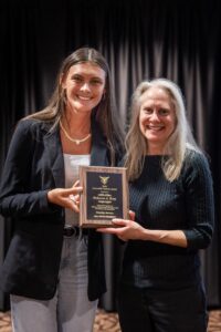Student Gracee Gibson, at left, with Dr. Becci Brey, at the Disability Awards Ceremony. They are both smiling, and posing for the camera holding the Accessible Teacher Award Plaque. Gracee nominated Dr. Brey for the award. They are both wearing black sweaters, and standing in front of a black velvet curtain background.