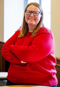 Portrait of Sarah Sager, in a red sweater. She is smiling, and looking at the camera. 