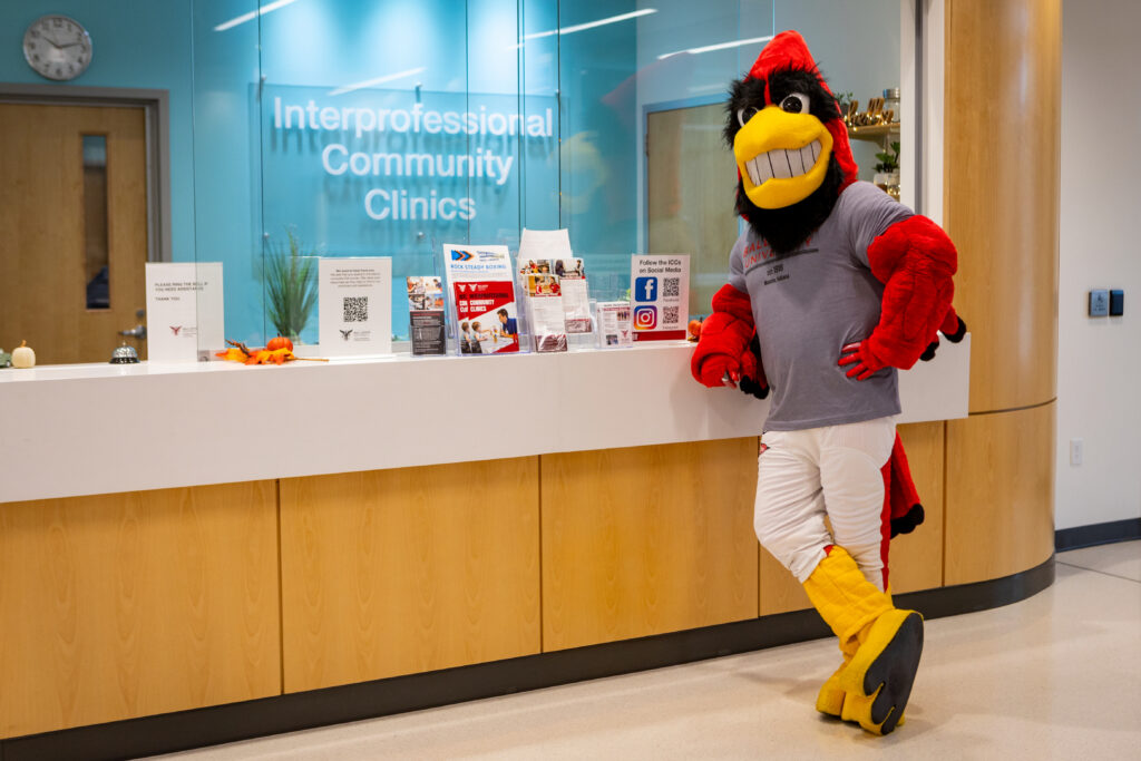 Charlie Cardinal is pictured on the right hand side of this photo, leaning on the front counter at the Interprofessional Community Clnic. The front desk is made of a blonde wood, with white countertop, and large glass sliding windows encasing the desk area. Behind Charlie you can see a big sign that says Interprfessional Community Clinic. It is white letters on glass, mounted on a teal blue painted wall. There are various other small signs/ pamphlet displays that are unreadable in the photo for clinic services and or other community partner services dotting the front counter.