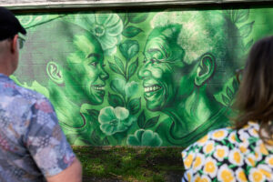 Two people with their backs to the camera stand framing a beautiful green-toned mural of an older person's face and a child's face smiling at each other across flower embellishments.