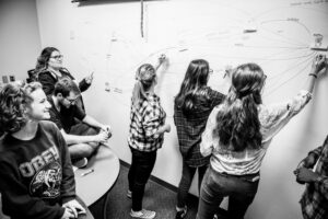 A group of people are in front of a whiteboard, some are writing on it while others watch